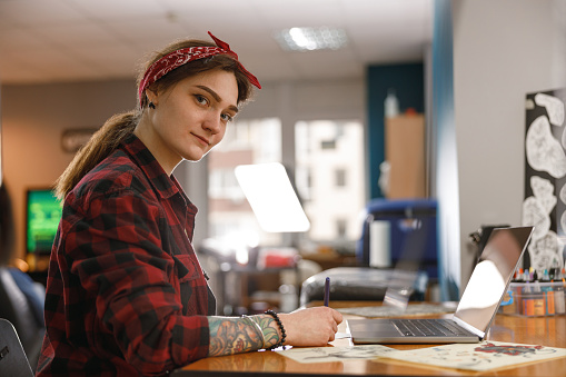 Portrait of attractive artist woman sitting at table in her office and drawing something, smiling at camera