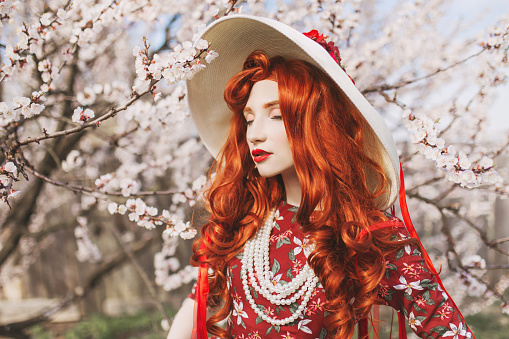 Woman with long red hair admiring blooming flowers on spring trees. Beautiful redhead girl enjoy simple things. Wellness and self-care. Flower smell. Vanilla dress. Cherry garden. Mental health