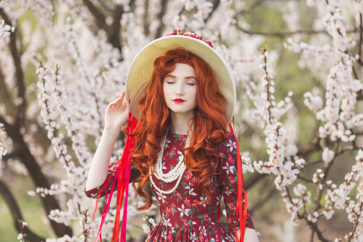 Woman with long red hair admiring blooming flowers on spring trees. Beautiful redhead girl enjoy simple things. Wellness and self-care. Flower smell. Vanilla dress. Cherry garden. Mental health