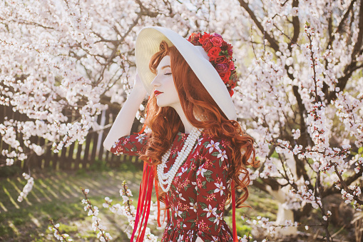 Woman with long red hair admiring blooming flowers on spring trees. Beautiful redhead girl in hat enjoy simple things. Wellness and self-care. Flower smell. Vanilla dress. Cherry garden. Mental health