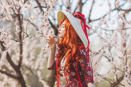 Woman with long red hair admiring blooming flowers on spring trees. Beautiful redhead girl enjoy simple things. Wellness and self-care. Flower smell. Vanilla dress. Cherry garden. Mental health