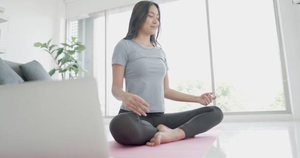 Young asian woman wear activewear sit on mat, practicing yoga in lotus position relaxing for stress relief in living room, follows an online exercise course video on laptop. Health, leisure activitie. joven asiática usa ropa deportiva siéntese en la colchoneta, practicando yoga en posición de loto relajándose para aliviar el estrés en la sala de estar, sigue un video del curso de ejercicios en línea en la computadora portátil. actividades de salu