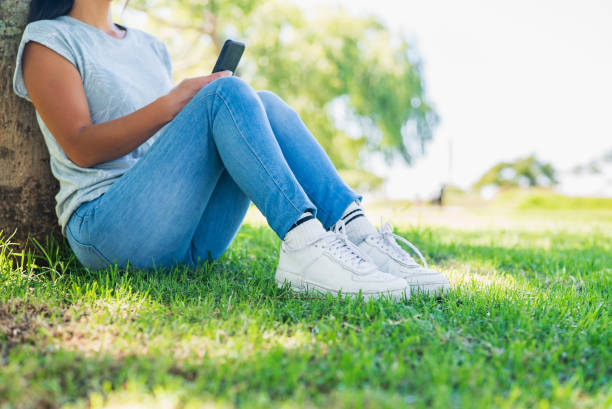 Low section of asian american woman sitting in park while leaning on tree using smart phone during daytime