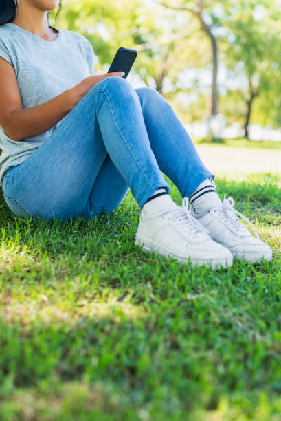 Low section of asian american woman sitting in park while leaning on tree using smart phone during daytime