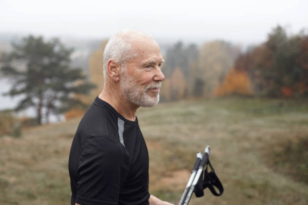 side view of strong athletic seventy year old caucasian in black dry fit t-shirt enjoying autumn morning in wild nature, posing in colorful foggy forest, holding nordic walking poles and smiling - 80 year old imagens e fotografias de stock