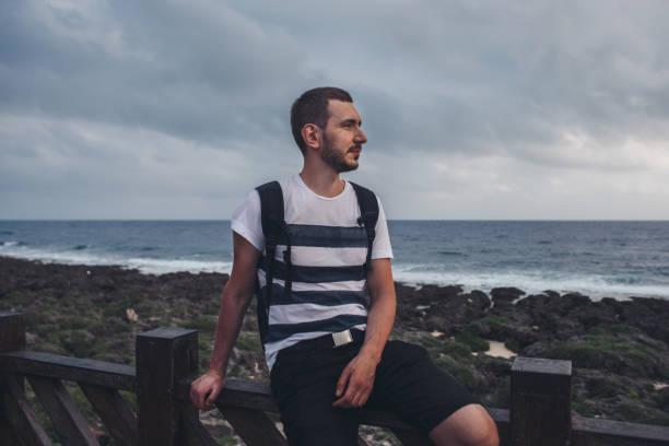 Young man sits on a railing by the sea.