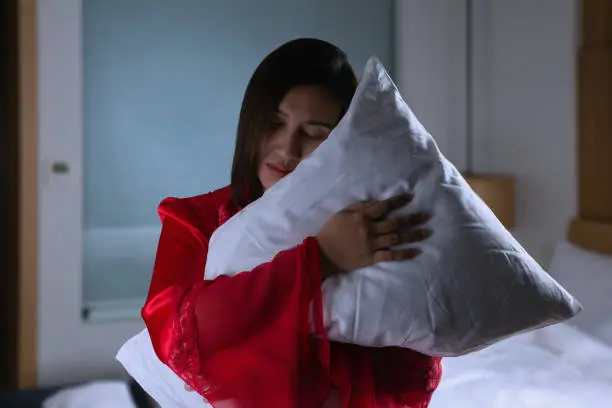 Asian woman in a silk red nightgown standing hugging pillow with two hands in a bedroom at night. Female in satin nightwear and embracing soft pillow