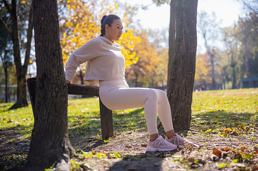 Asian woman in casual sports clothes exercising muscles and doing dips, leaning on log hurdle in the park, outdoor activities on sunny autumn day