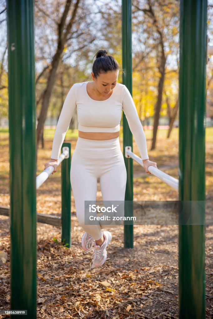 Asian woman in casual sports clothes working out on parallel bars in the park Asian woman in casual sports clothes exercising arm muscles on parallel bars in the park, bending legs in knee, leisure outdoor workout on sunny autumn day 35-39 Years Stock Photo