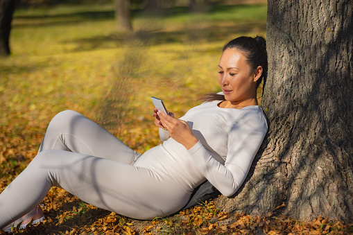 Asian woman in casual sports clothes sitting on dry leaves and leaning on a tree, texting on mobile phone and relaxing on sunshine