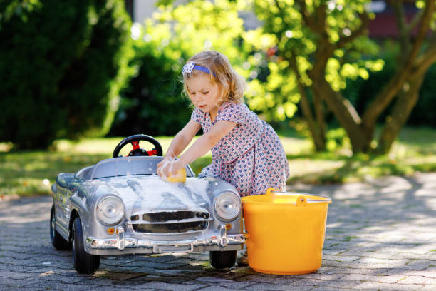 bonita hermosa niña niña niña lavando gran coche de juguete viejo en el jardín de verano, al aire libre. feliz niño sano limpiando el coche con agua y jabón, divirtiéndose con salpicaduras y jugando con la esponja. - outdoor toy fotografías e imágenes de stock