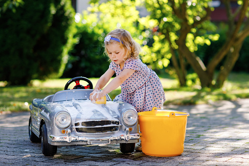 Cute gorgeous toddler girl washing big old toy car in summer garden, outdoors. Happy healthy little child cleaning car with soap and water, having fun with splashing and playing with sponge