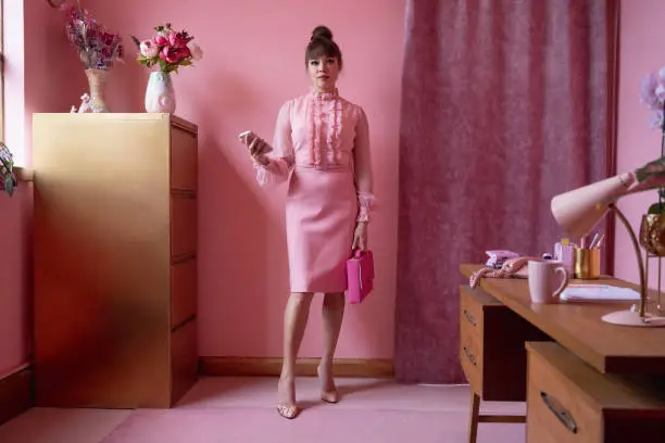 Full length front view of retro style businesswoman in early 40s wearing pink blouse and skirt and looking at camera while standing in matching pastel office.