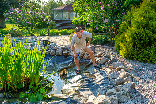 Young male in casual clothes looking at camera, while removing mud and sludge from bottom of artificial pond with landing net on sunny summer day in garden