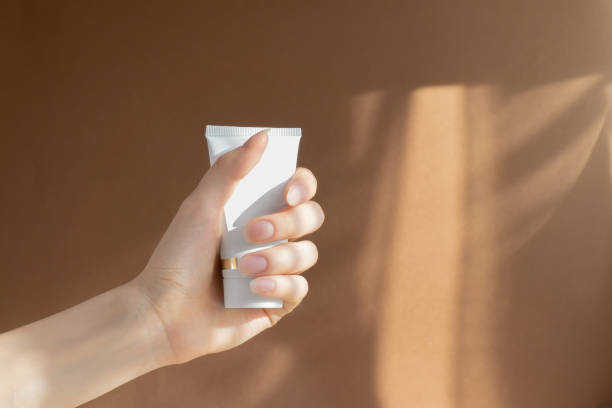 istock Woman's hand clutches white unmarked cream bottle in her palm. Container with body lotion, toiletries. Tube for professional care products on brown background in rays of sunlight 1385657505