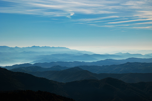 beautiful hills and mountains, colored of shades of blue, extending up to the horizon at daytime, while being lit by the sunlight, bright fog passing through the valleys, light blue sky filled with various thin little clouds