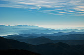 beautiful hills and mountains, colored of shades of blue, extending up to the horizon at daytime, while being lit by the sunlight, bright fog passing through the valleys, light blue sky filled with various thin little clouds