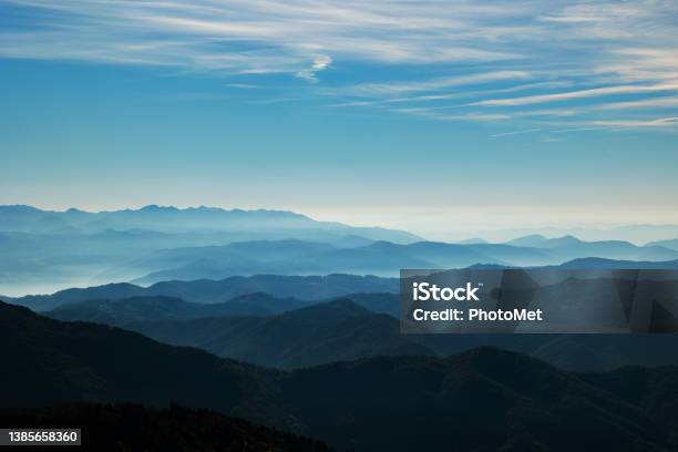 Beautiful Hills And Mountains Colored Of Shades Of Blue Extending Up To The Horizon At Daytime While Being Lit By The Sunlight Bright Fog Passing Through The Valleys Light Blue Sky Filled With Various Thin Little Clouds Stock Photo - Download Image Now