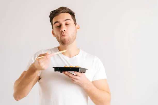 Photo of Portrait of handsome young man with enjoying eating fresh tasty sushi rolls with chopsticks on white isolated background.