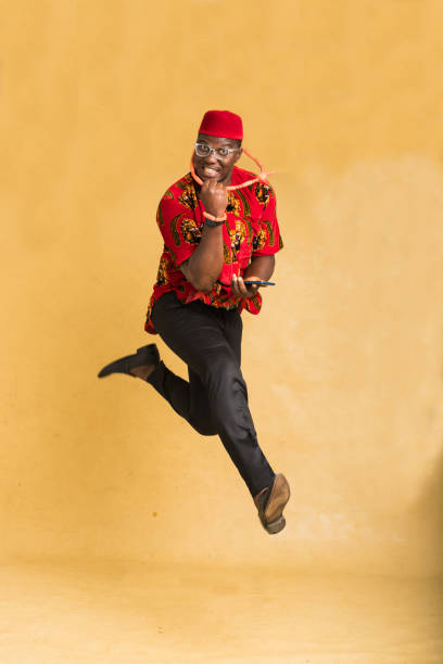 igbo traditionally dressed business man in mid air with phone in hand front view - nigeria african culture dress smiling imagens e fotografias de stock
