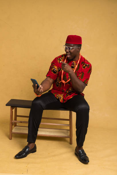 igbo habillé traditionnellement homme d’affaires assis et regardant le téléphone et joyeux - nigeria african culture dress smiling photos et images de collection