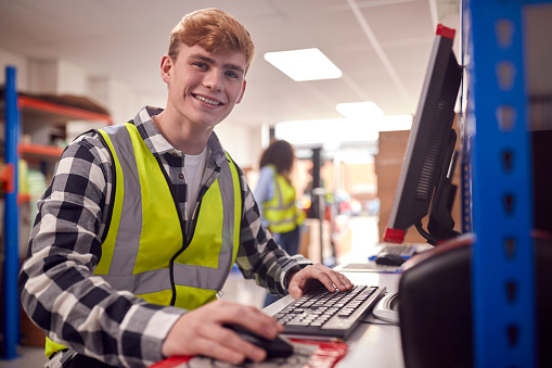 Portrait Of Male Intern Working In Busy Modern Warehouse On Computer Terminal