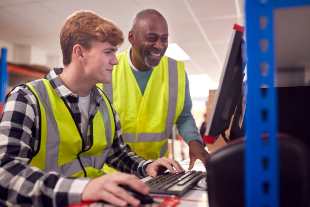 male intern with supervisor working in busy modern warehouse on computer terminal - trainee imagens e fotografias de stock