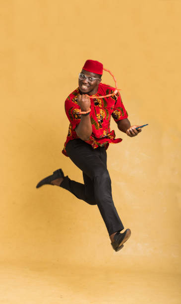 igbo traditionally dressed business man in mid air with phone in hand celebrating - nigeria african culture dress smiling imagens e fotografias de stock