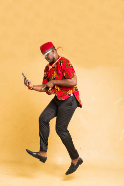igbo homme d’affaires habillé traditionnellement dans les airs avec téléphone à la main heureux - nigeria african culture dress smiling photos et images de collection