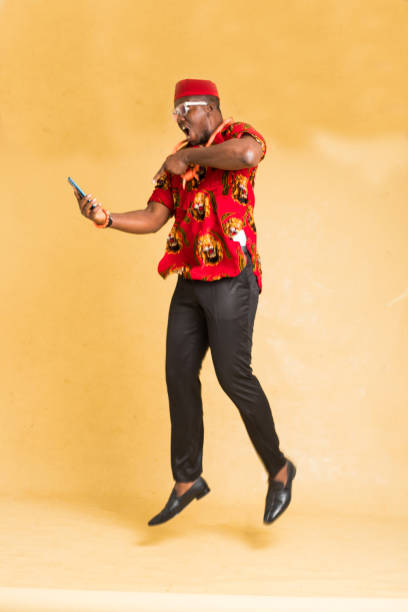 igbo traditionally dressed business man in mid air with phone in hand - nigeria african culture dress smiling imagens e fotografias de stock