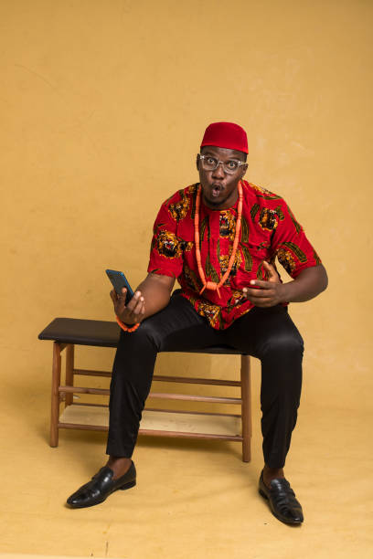 igbo tradycyjnie ubrany biznesmen siedzący, z telefonem i radosny - nigeria african culture dress smiling zdjęcia i obrazy z banku zdjęć