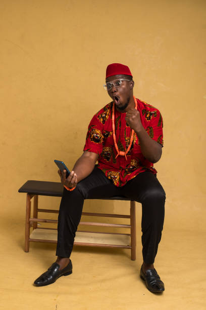 igbo homme d’affaires habillé traditionnellement assis et regardant le téléphone et la pompe à poing - nigeria african culture dress smiling photos et images de collection