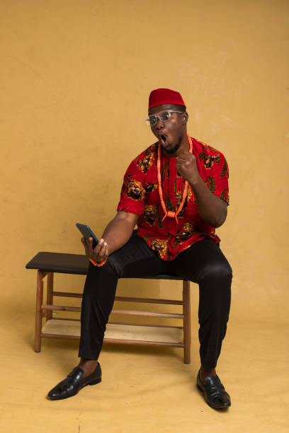 igbo tradicionalmente vestido homem de negócios sentado e olhando para telefone e bomba punho - indigenous culture nigerian culture african descent nigeria - fotografias e filmes do acervo