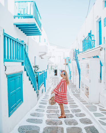 Back view of a young beautiful happy traveller female lady with a red dress and bag enjoys the scenery and walking through the traditional white and blue small, whitewashed alleys of Mikonos town, Cyclades islands of Greece during summertime.