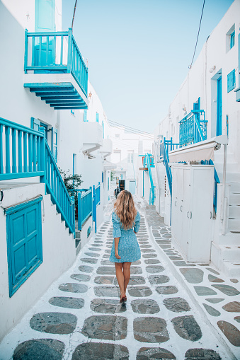 Back view of a young beautiful happy traveller female lady with a blue dress enjoys the scenery and walking through the traditional white and blue small, whitewashed alleys of Mikonos town, Cyclades islands of Greece during summertime.