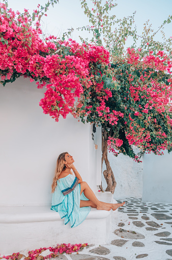 Back view of a young beautiful happy traveller female lady with a blue dress and bag enjoys under the red bougainvillaea or begonvil flower scenery and sitting and looking up through the traditional white and blue small, whitewashed alleys of Mikonos town, Cyclades islands of Greece during summertime.