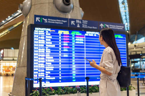 woman with luggage and smart phone checking flight schedule near departure board in airport.