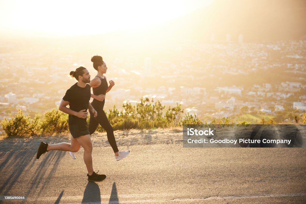 Two fit young people jogging together along a scenic road Two fit young friends in sportswear jogging together along a scenic road overlooking the city on a sunny afternoon Running Stock Photo
