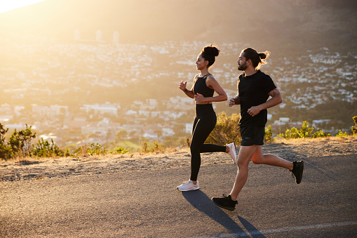 Two fit young friends in sportswear smiling while out for a run together along a scenic road overlooking the city on a sunny afternoon