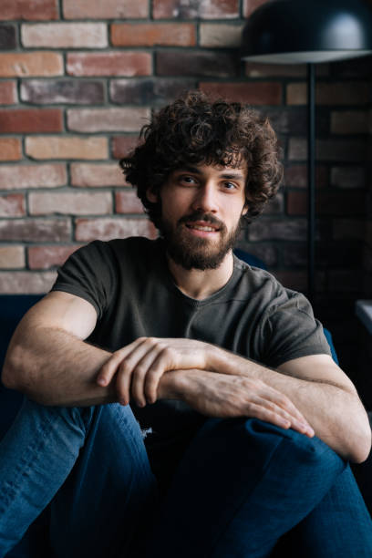 istock Vertical portrait of confident bearded young man smiling looking at camera while sitting on comfortable sofa in living room with brick wall. 1385691817