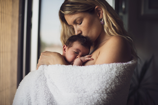 Newborn baby in mother's arms. Beautiful young mother is holding her newborn baby girl wrapped in blanket while sleeping. Baby girl feels love.