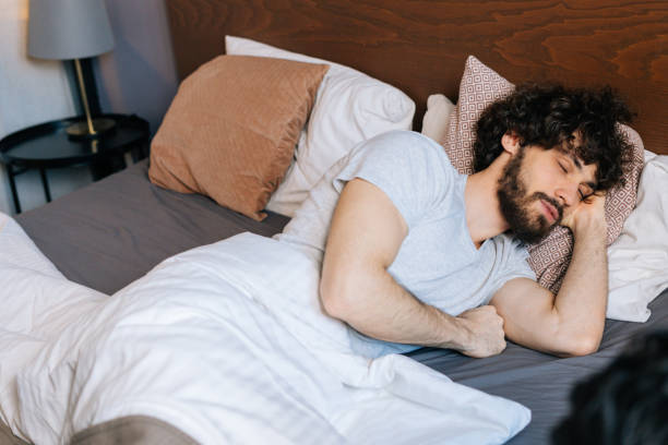istock High-angle view of tired bearded young man sleeping peacefully lying on side in large comfortable double bed under white blanket with head on pillow. 1385697679