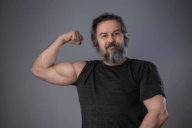 istock Bearded senior man man showing biceps on gray wall background. 1385705460