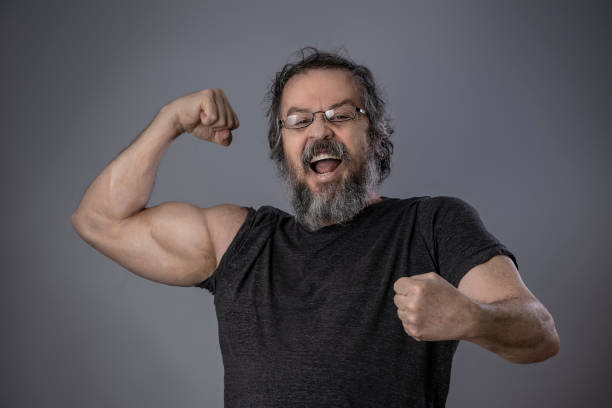 istock Bearded senior man man showing biceps on gray wall background. 1385705519