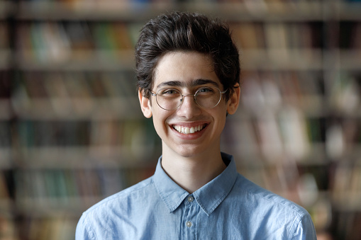Head shot portrait of happy handsome Jewish male student in eyeglasses posing at blurred library background. Smiling Z generation smart guy looking at camera, high school or college education concept.