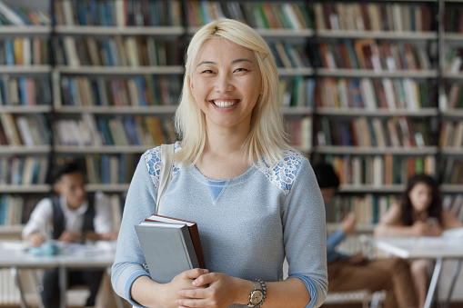 Portrait of happy young smart Asian female student with books in hands posing in modern public or college library. Smiling beautiful millennial young Japanese girl looking at camera, education concept