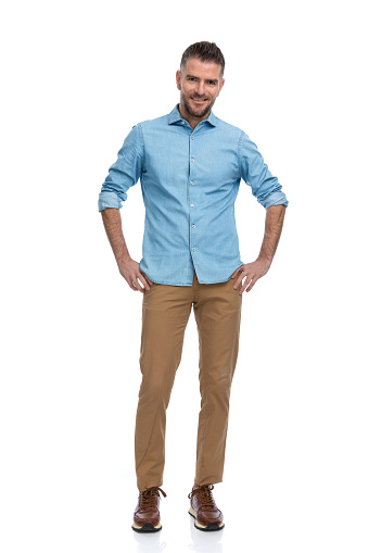 full body picture of handsome unshaved guy with chino pants and sneakers smiling while holding hands on hips in front of white background