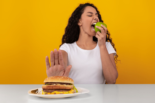 Say No To Junk Food. Lady Refusing To Eat Unhealthy Burger Choosing Biting Apple, Showing Stop Sign Gesture With Palm Hand To Hamburger Sitting At Table, Selective Focus, Yellow Orange Studio Wall