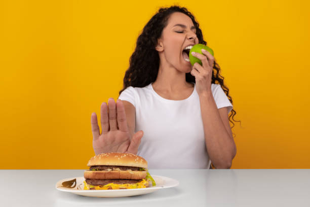Portrait of Pensive Lady Holding Apple And Burger Say No To Junk Food. Lady Refusing To Eat Unhealthy Burger Choosing Biting Apple, Showing Stop Sign Gesture With Palm Hand To Hamburger Sitting At Table, Selective Focus, Yellow Orange Studio Wall avoidance stock pictures, royalty-free photos & images