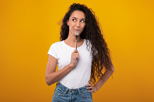 Portrait of excited hungry young woman holding fork in her mouth, biting and dreaming about delicious healthy food, choosing something to eat posing looking aside isolated on yellow orange studio wall